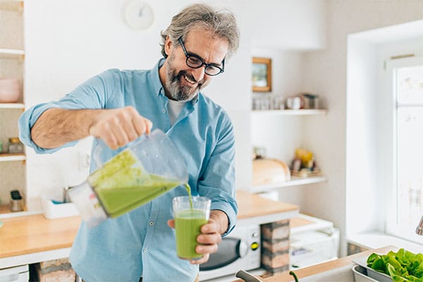 Gezeigt wird ein Mann in der Küche der sich einen gesunden grünen Smoothie einschenkt.