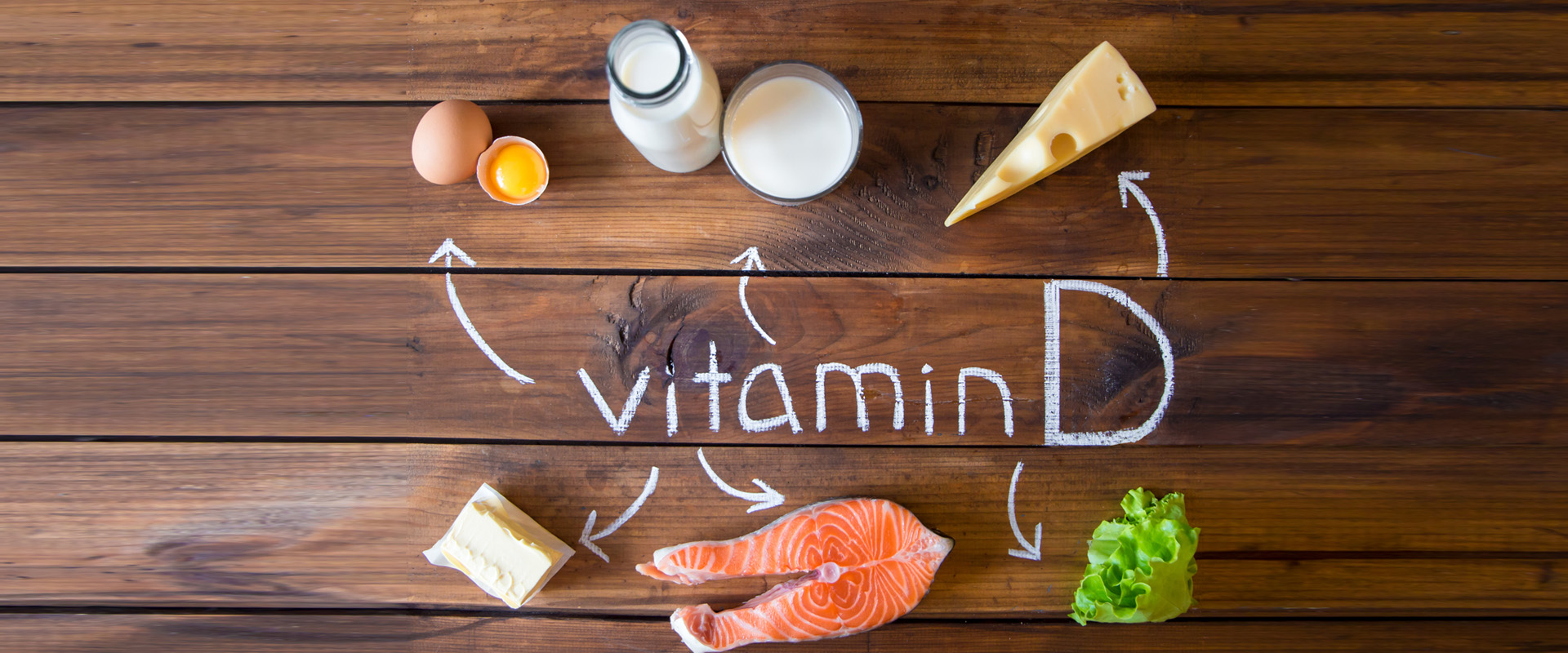 Lebensmittel mit Vitamin D wie Ei, Milch, Käse, Butter, Fisch und grüner Salat liegen auf einem Holztisch