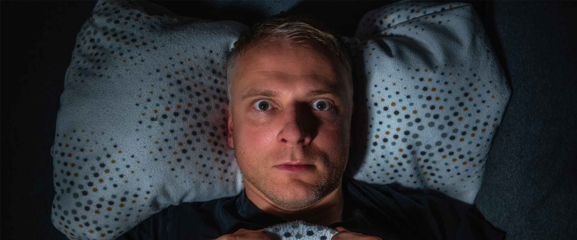 Ein Mann liegt verzweifelt mit weit aufgerissen Augen wach im Bett, klammert sich an seine Bettdecke und kann nicht einschlafen