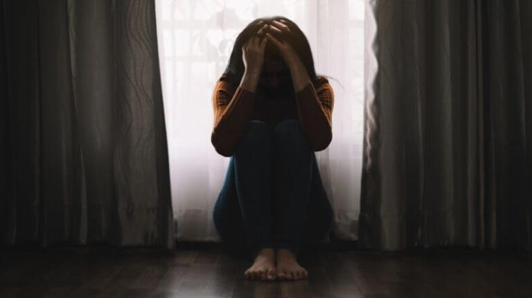 Eine Frau kauert in vor einem Fenster in einem dunklen Raum und wirkt traurig und depressiv.